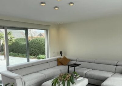 Moderne woonkamer met lichte hoekzetel, grote ramen en strak geschilderde muren en plafonds.
